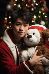 Fototapeta na wymiar handsome and cute young guy sitting near the Christmas tree and hugging a teddy bear in a santa claus costume