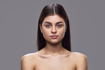 Frontal portrait of young woman with clear skin, natural makeup, and neat hair against gray background. Concept of natural beauty and serenity, skin care products, nutritional supplements.