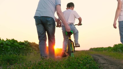 Father grasps bicycle seat providing support to kid pedaling by mother on plantation. Mother gazes...