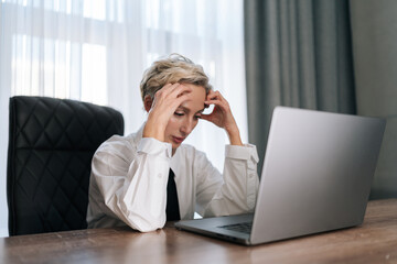 Exhausted upset middle-aged businesswoman sitting at table with laptop, leaning head on hand,...