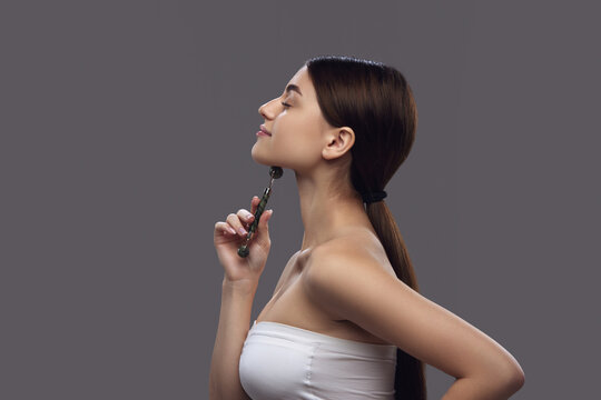 Side view portrait of young woman using roller from natural stone, to smooth wrinkles and prevent double chins. Concept of facial treatment and daily skincare routines using eco-friendly tools.