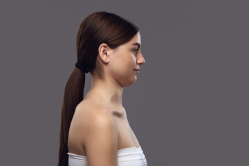 Side profile of young brunette woman with ponytail and bare shoulders, doing exercises for prevent double chin against neutral background. Concept of facial care and beauty treatment.