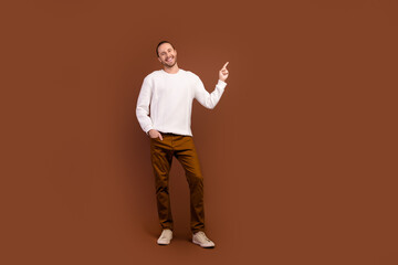 Fototapeta na wymiar Full length photo of funky cheerful guy dressed white pullover pointing finger empty space isolated brown color background