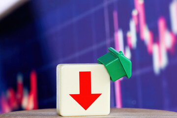 The concept of decline in the real estate market is illustrated with a plastic green house falling down, a plastic block inscribed with red arrow and a red graph in the background.