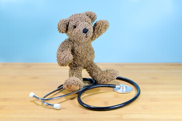 Little brown teddy bear standing on a stethoscope and waving, health care for children, pediatric medicine concept, blue background with copy space - obrazy, fototapety, plakaty
