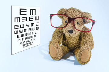 Little teddy bear with big red glasses sitting beside a pediatric vision screening chart, concept of eye care and ophthalmology for children, light blue background, copy space
