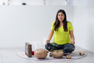 A woman meditating in a white room, sitting on the floor with bowls around her