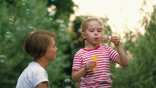 Caring mother looks at child focused on blowing bubbles in fresh air during holidays. Schoolgirl stands next to mother blowing bubbles. Daughter spends time with mom holding soap bubbles bottle