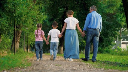 Happy kids stroll clasping hands with lovely parents in park on vacation. Elated parents relish meandering with interconnected hands near kids. Boy and girl amble hand in hand accompanied by parents