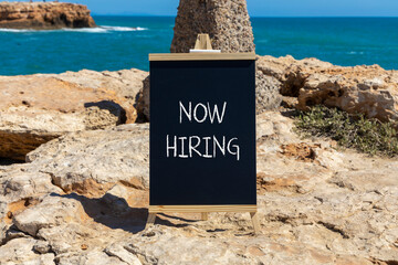 Now hiring symbol. Concept words Now hiring on beautiful black chalk blackboard. Beautiful stone sea blue sky background. Business marketing, motivational now hiring concept. Copy space.