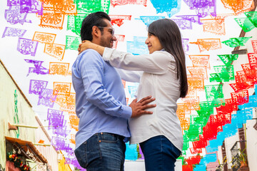 From below of couple lovingly embraces under a canopy of colorful papel picado celebrating their pregnancy in the festive streets of Tequisquiapan Queretaro Mexico