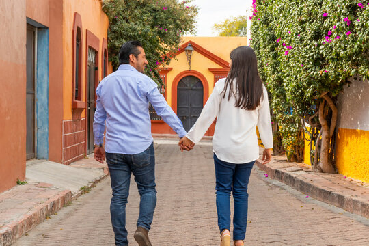 Back view of couple walks hand in hand along a colorful street in Tequisquiapan Queretaro Mexico surrounded by vibrant walls and blooming flowers