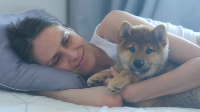 Happy young woman hugs cute puppy shiba inu dog lying on bed together at home interior. Close up. Young people, domestic animals, concept of happiness, friendship and love between an animal and human