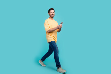 Full length photo of young brunet blogger start his career in social medias guy strolling with cellphone isolated on cyan color background