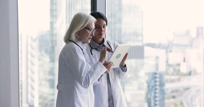 Younger intern woman consulting senior female doctor for expertise, diagnosis, showing online content on tablet, listening to advice. Two medical colleagues using digital gadget together