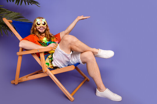 Full size photo of cheerful man in sunglass sit on armchair with cocktail pulm hold offer empty space isolated on purple color background