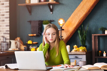 Young pensive woman freelancer sitting at table at home with laptop, making notes