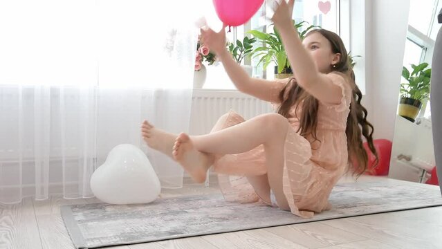 A little girl in a peach dress is sitting on the floor among balloons, joyfully tossing a red heart-shaped top. Valentine's Day is happiness, celebration.