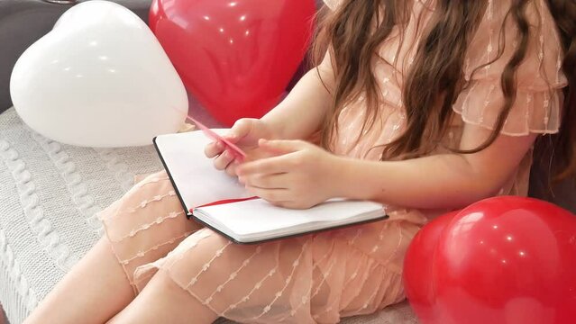 A little girl sitting on the couch opens a notebook book and picks up a bookmark or a postcard in the shape of a delicate pink heart. Valentine's Day, a lover's gift. first love.
