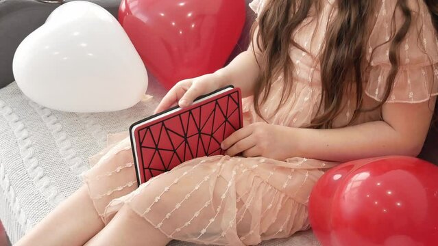 The hands of a little girl holding a red notebook diary sitting on the couch next to red and white balloons. Valentine's Day, a lover's gift. first love.
