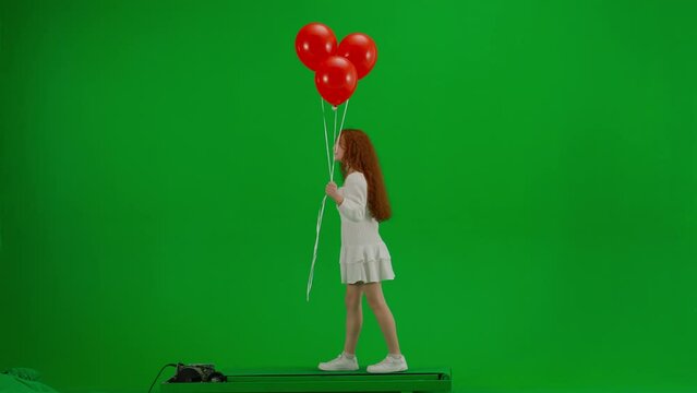 Redheaded little girl in white dress walking with red balloons on green studio background. Side view. Concept of holiday, joy and fun.
