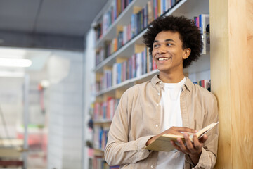 Happy black student guy holding book, looks away at free space, and smiling, leaning at shelf in...