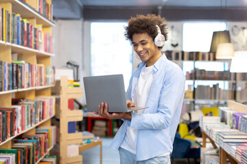 Happy black student guy standing in library, wearing headphones, holding laptop, radiating enthusiasm for tech-driven learning, and smiling