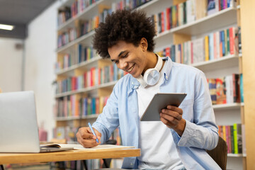 Black male student sitting in library, in front of laptop, using tablet, and writing notes, embodying multi-tasking approach to focused and productive study sessions