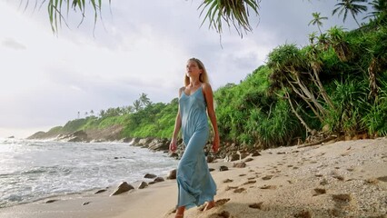 Elegant woman in blue dress strolls on sandy beach. Fashion model walks by sea, tropical backdrop. Luxury travel lifestyle, serene holiday. High-end resort wear display, summer collection showcase.