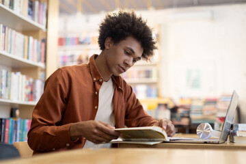 Focused African American student guy sitting at desk in university library, reading book, preparing for an exam, dedicated scene of academic focus and preparation