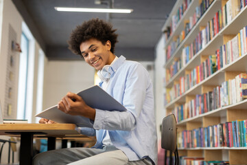 Smiling black male student sitting in the library, writing notes in copybook, holding book,...