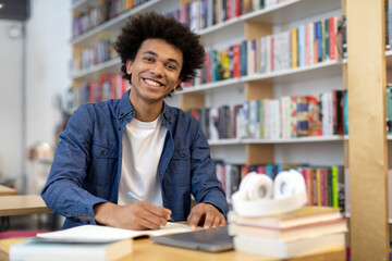 Happy black student guy sits at desk in library, smiling at the camera while writing in copybook,...