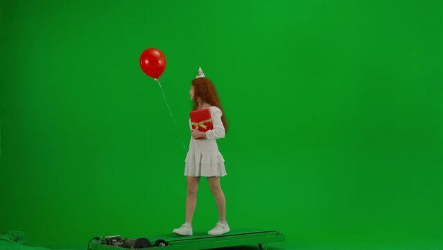 Redheaded little girl in white dress with a festive cap on her head and with a gift in her hands is walking with a red balloon. Green screen. Half turn. Concept of holiday, joy and fun.