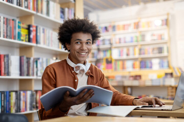Happy black student guy holding copybook and using laptop in university library, smiling at camera,...