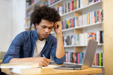 Sad, tired black student guy writing in copybook, sitting in front of laptop in college library,...
