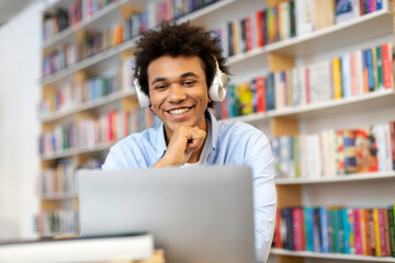 Happy black male student sitting at library desk, in front of laptop, wearing headphones and...