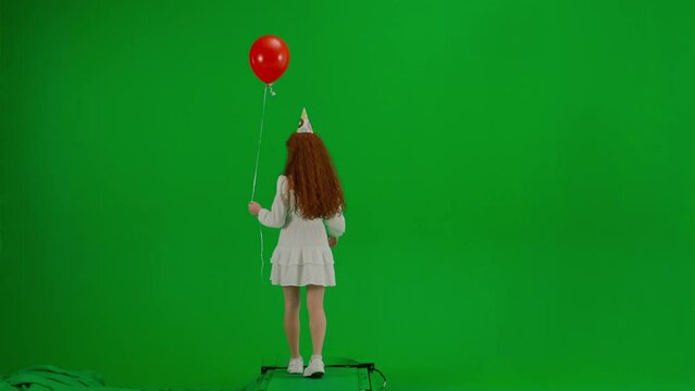 Redheaded little girl in white dress and with holiday cap on her head walking with red balloons on green background of studio. Back view. Concept of holiday, joy and fun.