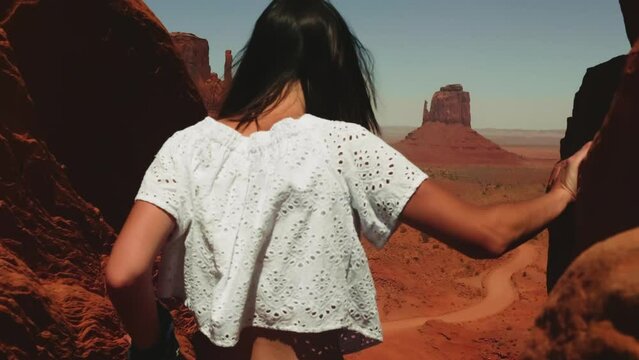 Back view young happy tourist woman walks between two rocks to epic desert vista point at Monuments Valley arms open.