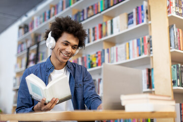 Multitasking black student guy sitting at library desk in front of laptop, wearing headphones,...