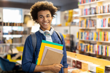 Cheerful african american male student, holds copybooks in library, backpack slung over his...