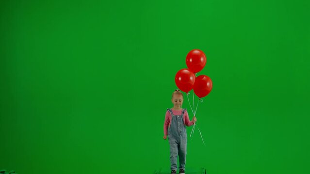 Little girl in jeans overall and ponytail with red helium balloons walking on chroma key green screen isolated background, front shot view.