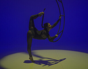 Fototapeta premium Young female aerial gymnast posing near moon on blue yellow studio background. Acrobat in black leotard shows stretching and holds her braid with her foot. Concept of aerial gymnastics, circus show.