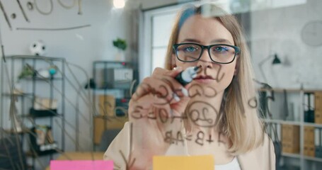 Attractive woman in glasses writing mathematical calculations on glass board in modern office,creativity concept - Powered by Adobe