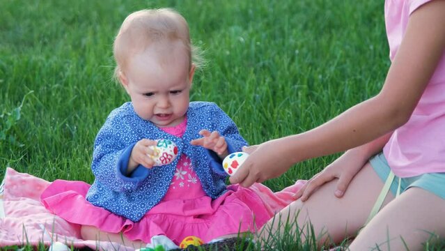 Little girl in bunny ears with her cute baby sister tapping with eggs while resting a on grass after Easter hunt