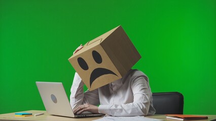 Woman in cardboard box with negative emoji on her head on green studio background. A worker sits at a desk, typing on a laptop, tired and frustrated.