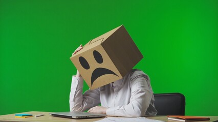 Woman in cardboard box with negative emoji on her head on green studio background. Tired, frustrated worker sitting at desk holding her head. Business life and daily routine in office.