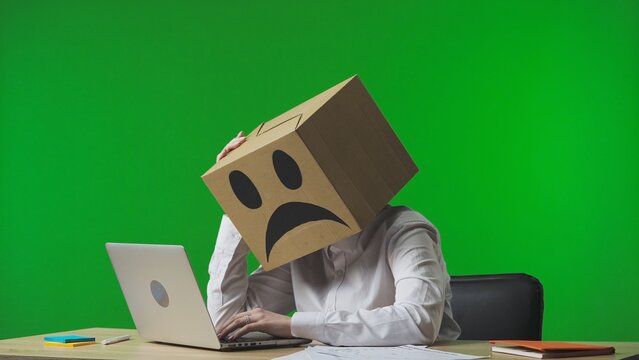 Woman in cardboard box with negative emoji on her head on green studio background. A worker sits at a desk, typing on a laptop, tired and frustrated.