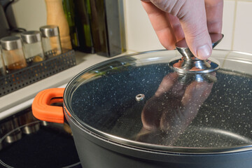 A hand holds the handle of the lid of a large cauldron on the stove, close-up