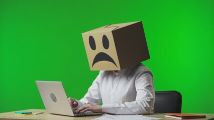 Woman in cardboard box with negative emoji on her head on studio green background. Worker typing on laptop keyboard, very upset and tired. Business life and daily routine in office.