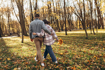 Heterosexual caucasian young loving couple walking outside in the city park in sunny weather, hugging smiling kissing laughing spending time together. Autumn, fall season, orange yellow red maple leav